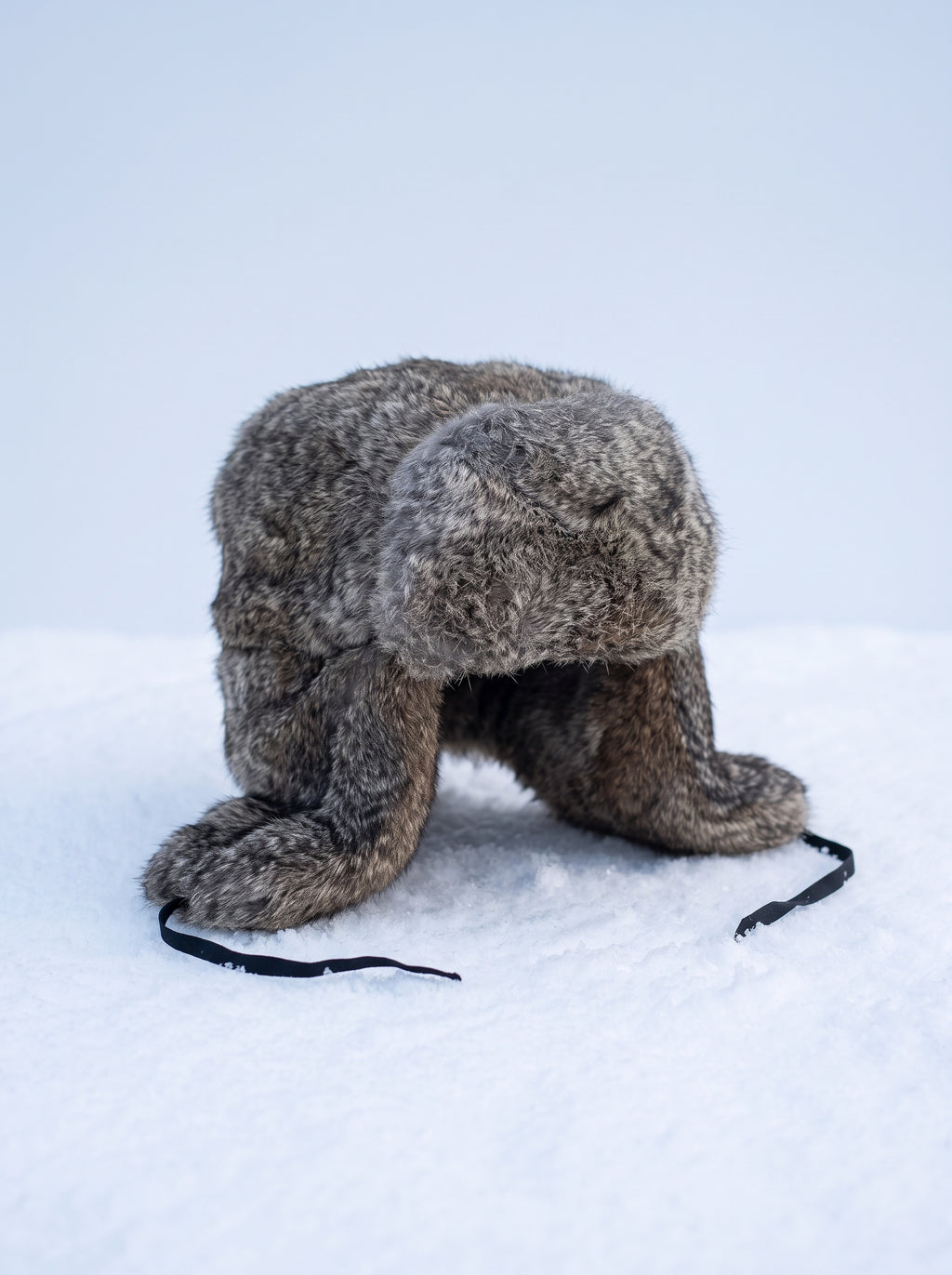 Chapka en fourrure de lapin grise, rabats baissés et sangles noires, posée sur la neige